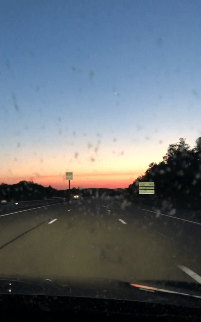 Vue du siège passager sur une autoroute au soleil couchant. Un panneau routier à droite et un autre plus loin. Il n'y a pas de voiture devant, mais les phares d'une voiture sont visibles sur la voie d'en face.