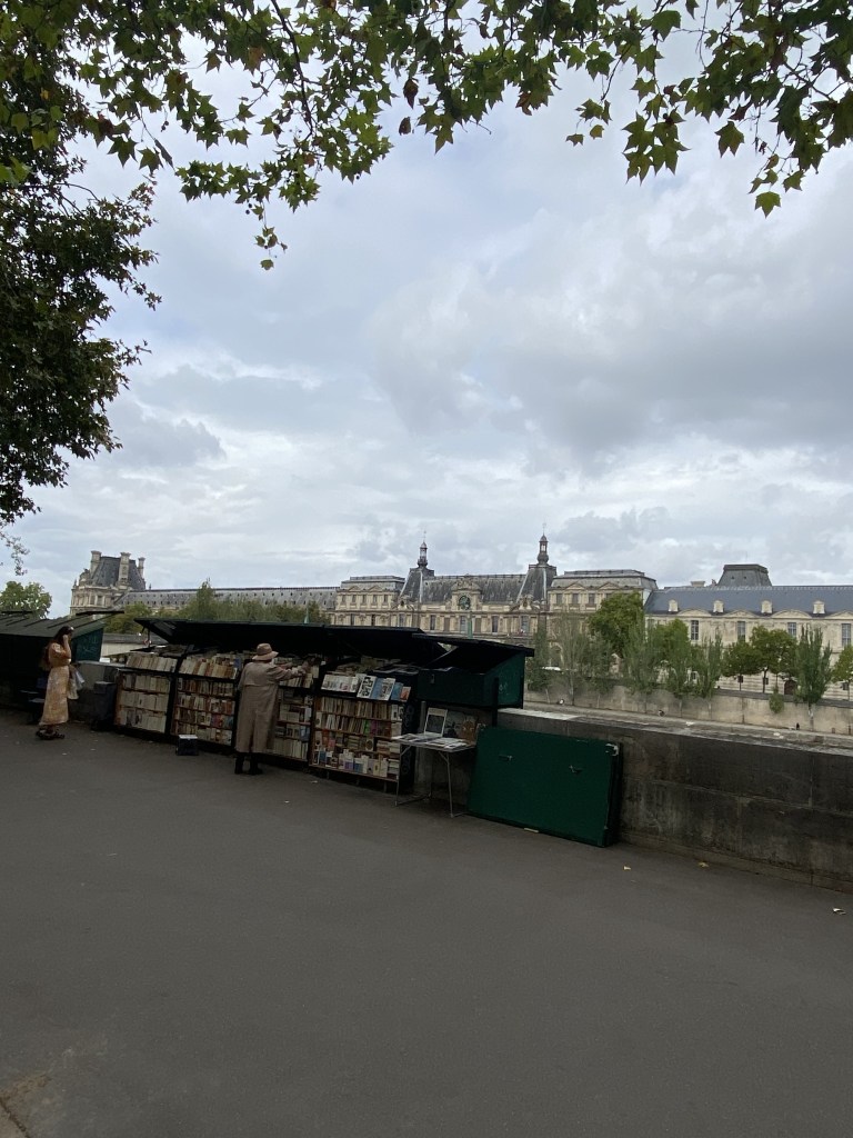 Les tuochants bouquinistes des Quais de Seine, ici Quai Voltaire rive gauche. En arrière plman de l'autre côté de la Seine, rive droite, on aperçoit le Louvre. Le ciel est un peu chargé.