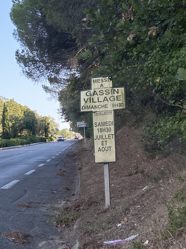 Un panneau en forme de croix latine, sur le bas-côté de la route indique les horaires de messe. Il est placé à l'ombre d'une généreuse verdure.