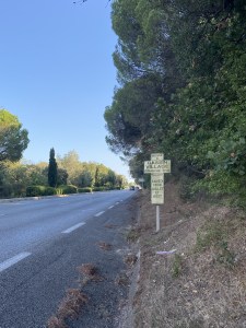 Un panneau en forme de croix, uur le bas-côté de la route. Il annonce les horaires de messe à l'entrée du village. le ciel estival est bleu et des beaux arbres méridionaux bordent la route