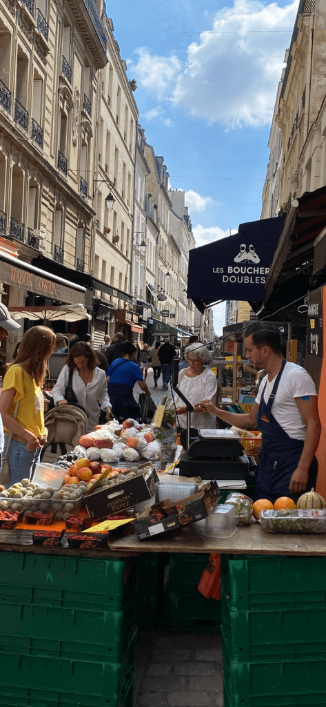Une jolie rue encaissée servant de marché de plein air. Un vendeur de primeurs sert une des nombtreuses clientes, sous un beau ciel bleu.