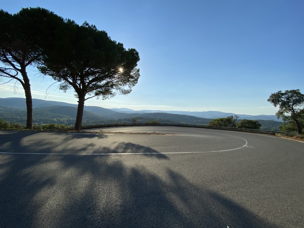 Belle route en épingle à cheveux. un grand pin parasol et des ombres d'arbres décorent cette route. Un beau ciel tout bleu sur fond de massif lointain.