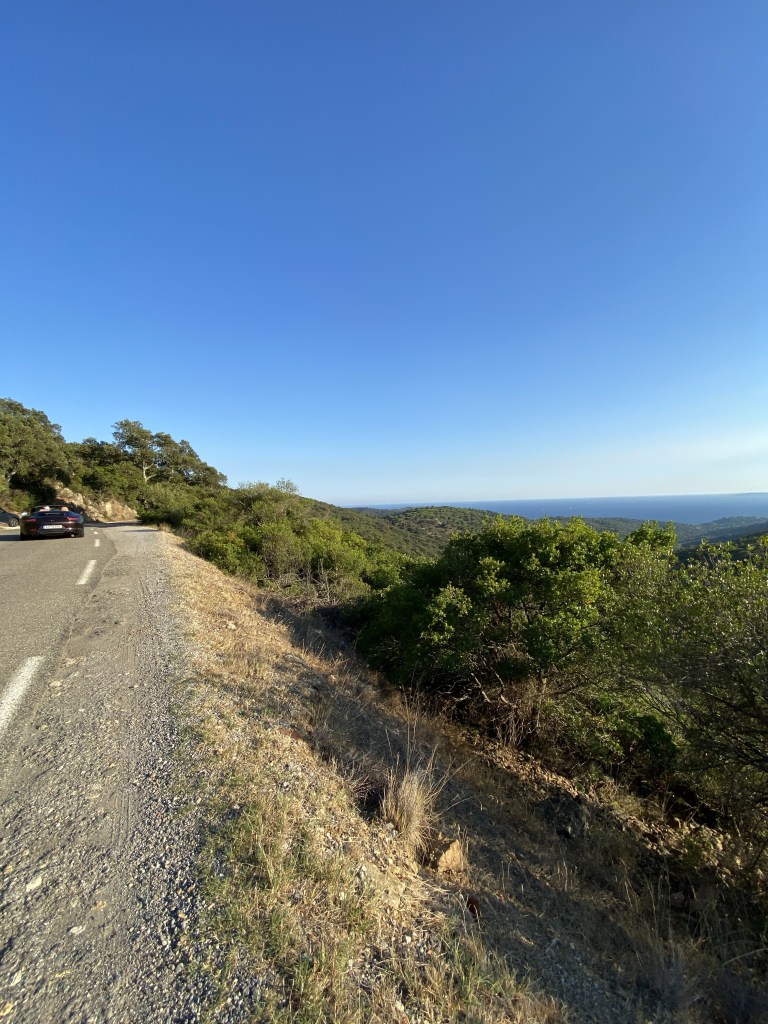 Une Porsche Carrera décapotée s'éloigne par une route touristique surplombant les versants verdoyants jusqu'à la Méditerranée, en contrebas .