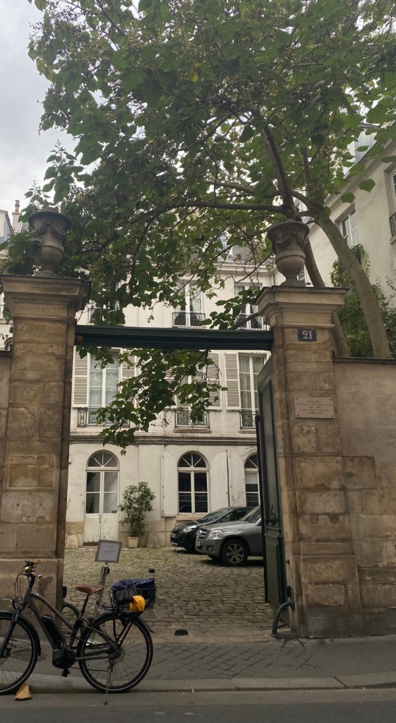 Un vélo est garé devant une majestueuse porte cochère ouvrant sur la cour d'un hotel particulier. Un arbre couronne de ses branchages cette entrée cochère.