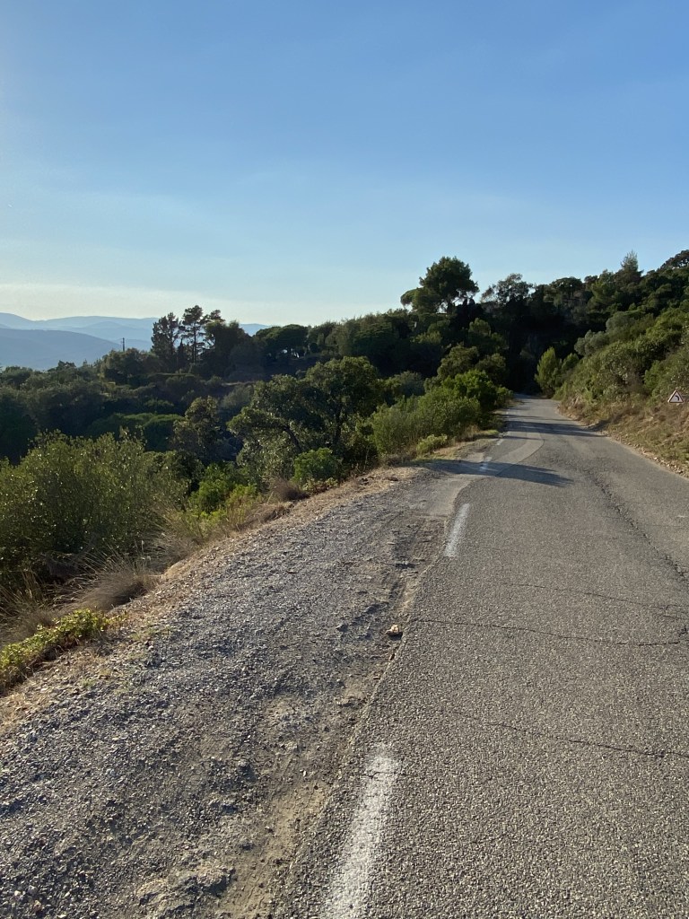 Vue d'une jolie route bordée de verdure épaisse, sous un ciel bleu d'été, dans le midi. Au fond on aperçoit le lointain massif des Maures.