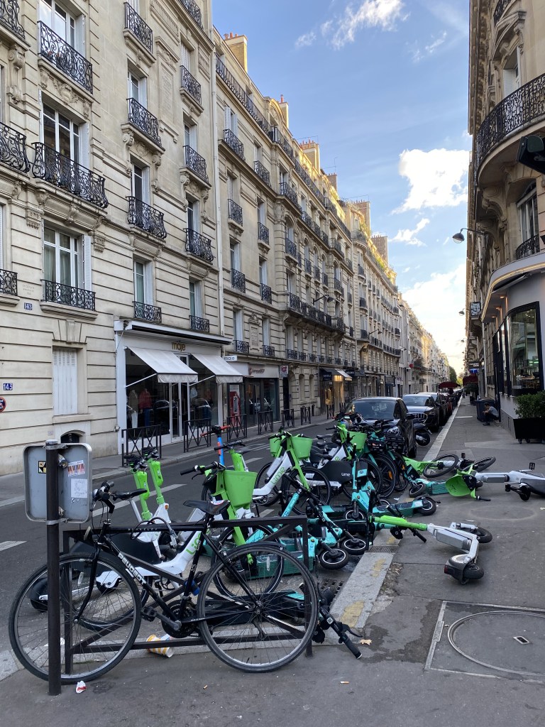 Des trottinettes garées en vrac, certaines se chevauchant et jonchant le sol. L'impression de grande négligence et de désordre est totale. Dommage, le ciel est bleu et la rue de la Pompe est bien rectiligne.