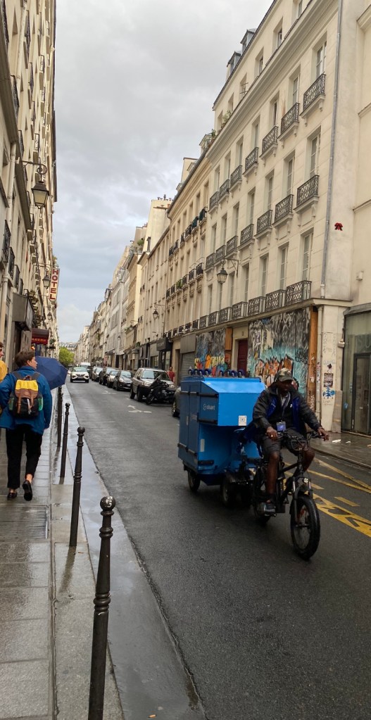 Un vélo cargo est manoeuvré par un livreur rue Notre Dame de Nazareth à Paris. La remorque est bleu, le ciel est couvert, une légère pellicule d'eau recouvre la chaussée après un petit passage pluvieux
