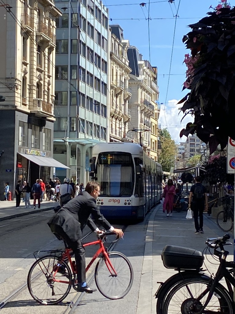 Un cycliste sur un vélo rouge traverse une rue passante, juste après le passage d'un tramway, rue de la Croix d'Or à Genève